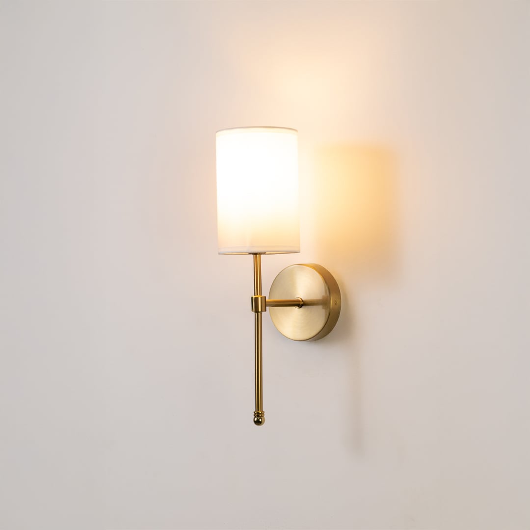 Metal gold wall light with white fabric shade glowing softly on a clean white background ideal for bedroom or hallway wall lighting.