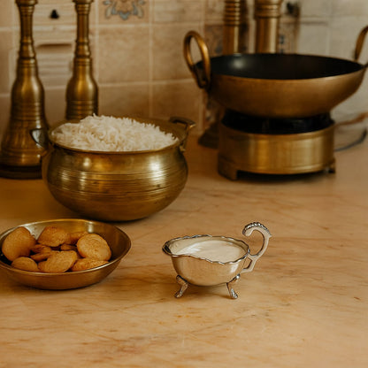 Elegant silver plated sauce bowl filled with white cream placed on a marble counter next to traditional brass cookware and rice handi.