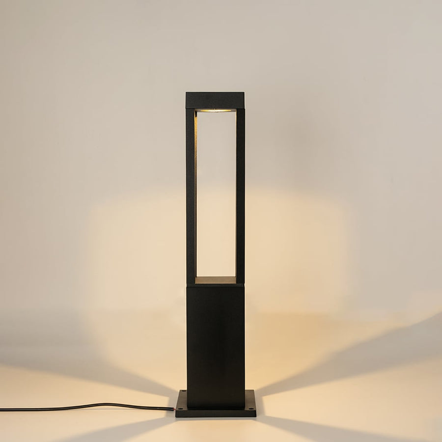 A contemporary black bollard light against a white background with the light on creating a warm focused glow that accentuates its minimal open frame structure.