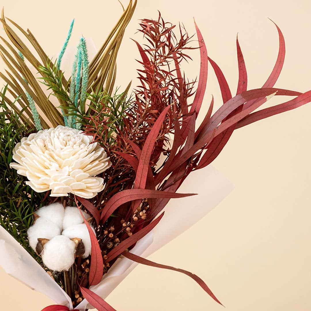 Closeup of dried bouquet with shola flower cotton stem palm leaf and red eucalyptus wrapped in white paper with satin ribbon.