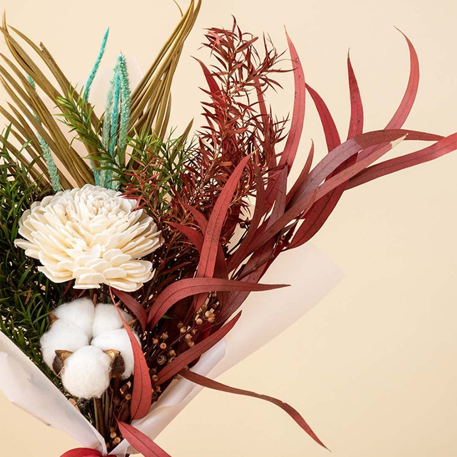 Closeup of dried bouquet with shola flower cotton stem palm leaf and red eucalyptus wrapped in white paper with satin ribbon.