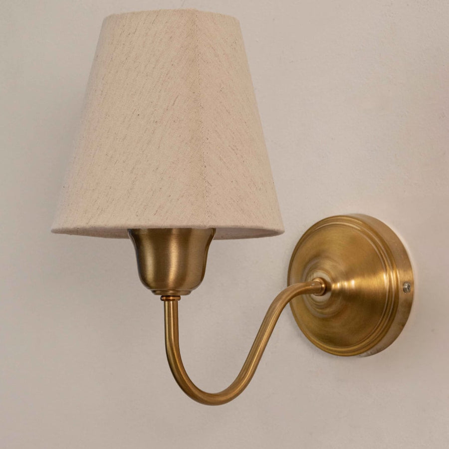 Close up view of brass wall sconce showing curved metal arm round decorative backplate and white fabric lampshade details.