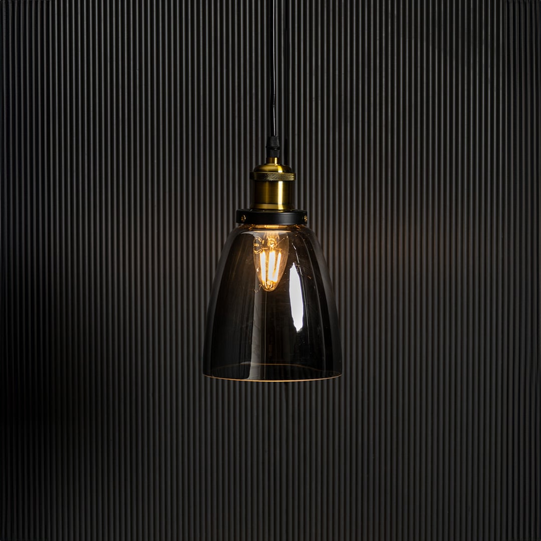 Smoked glass pendant light with brass and black holder turned on glowing softly against plain black background.