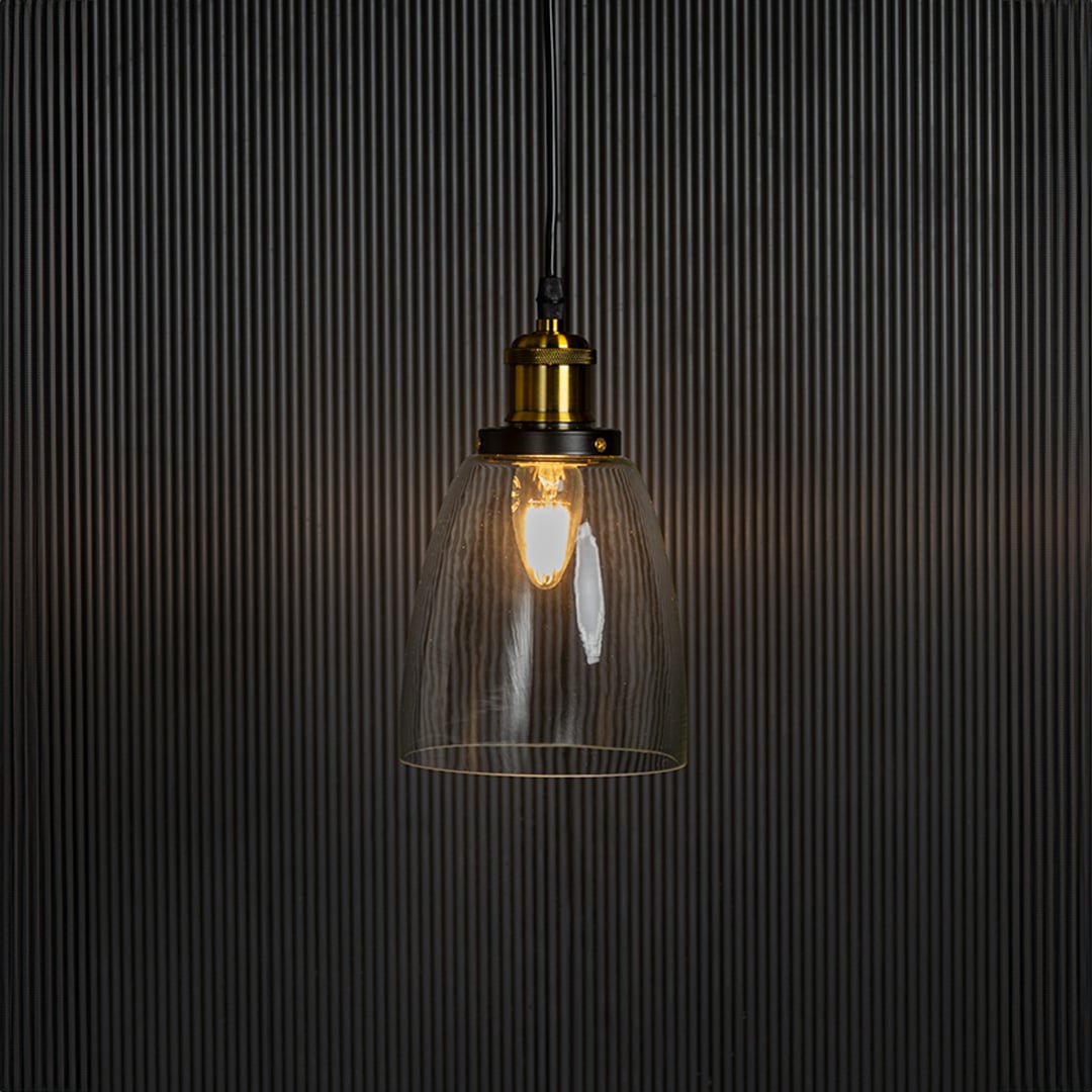 Clear glass pendant light with brass and black holder turned on glowing softly against plain black background.