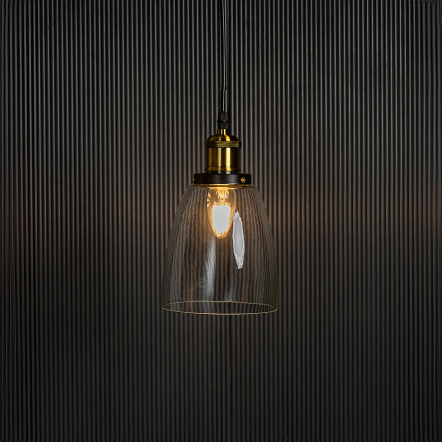 Clear glass pendant light with brass and black holder turned on glowing softly against plain black background.