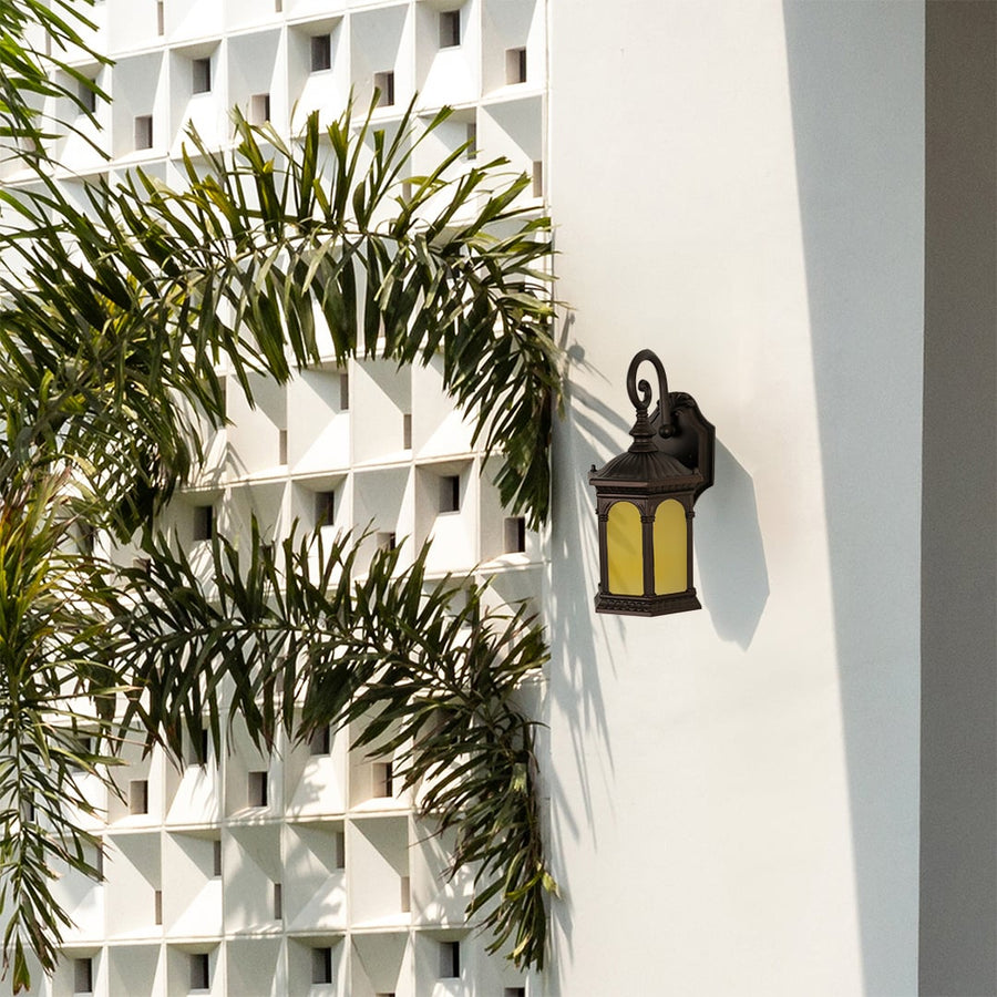 Large black traditional lantern wall lamp with textured glass illuminated on a white exterior wall next to palm trees and a decorative wall.