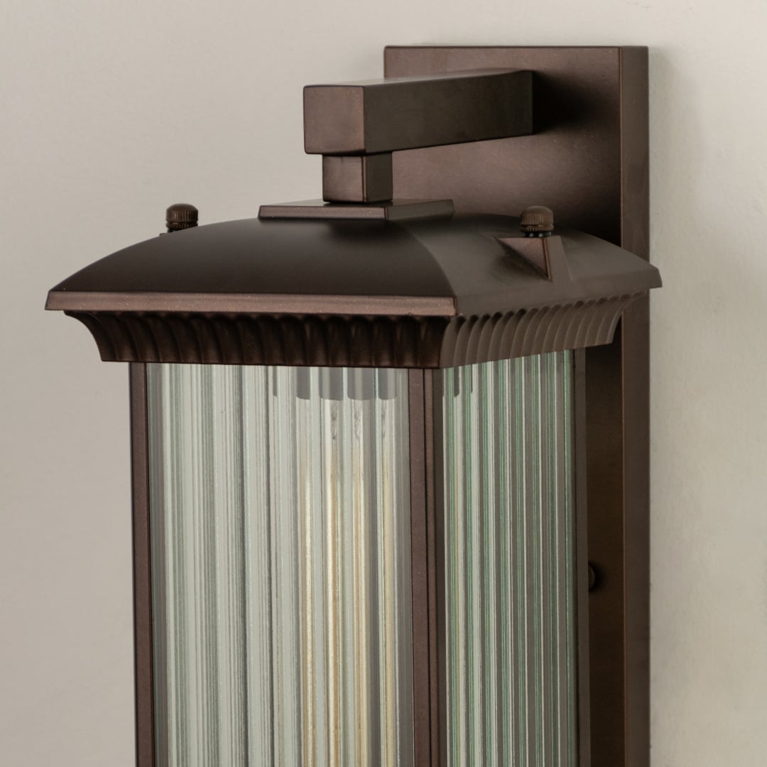 A closeup view of the bronze outdoor wall lantern showing the ribbed glass panels and warm glow highlighting the refined craftsmanship and elegant finish.