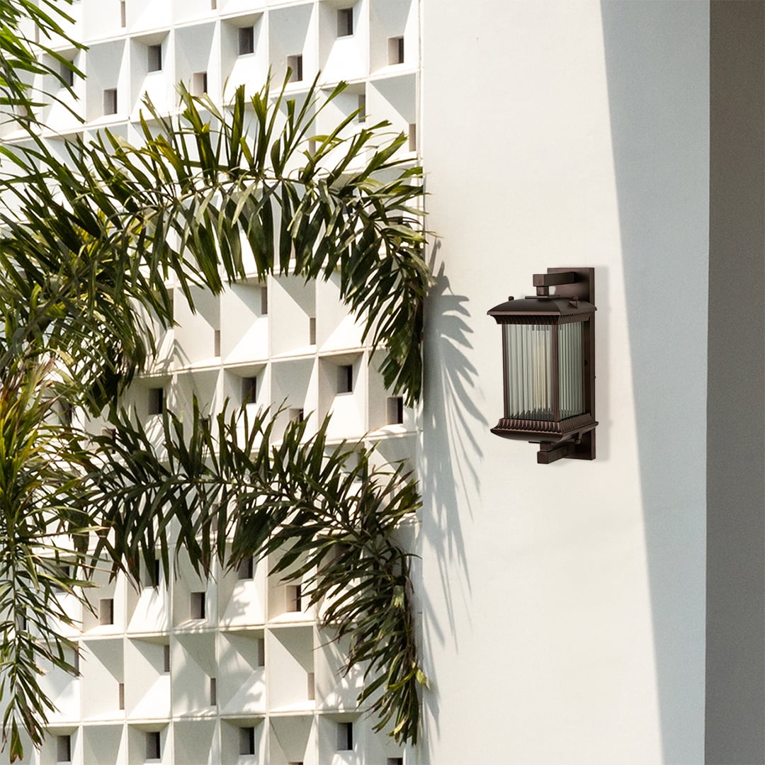 A stylish bronze outdoor lantern fixed on a white architectural wall glowing softly and adding character and charm to the surrounding outdoor environment.