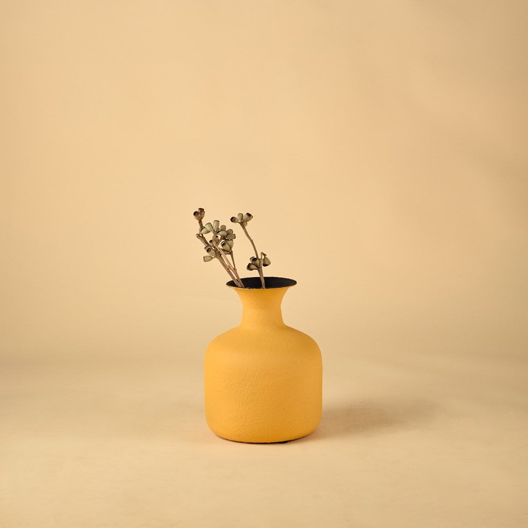A yellow textured vase with dried stems placed on a wooden dresser near a beige cushion and floral painting adding a calm warm and welcoming touch to the living space.