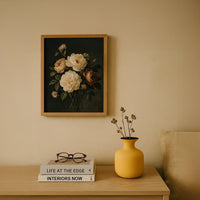 A yellow matte decorative vase displayed on a wooden surface beside stacked books and wall art in a softly lit interior setting creating a warm homely atmosphere.