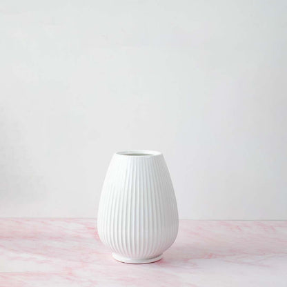 White ribbed ceramic vase with natural lighting on white background creating a soft shadow and minimalist artistic vibe.