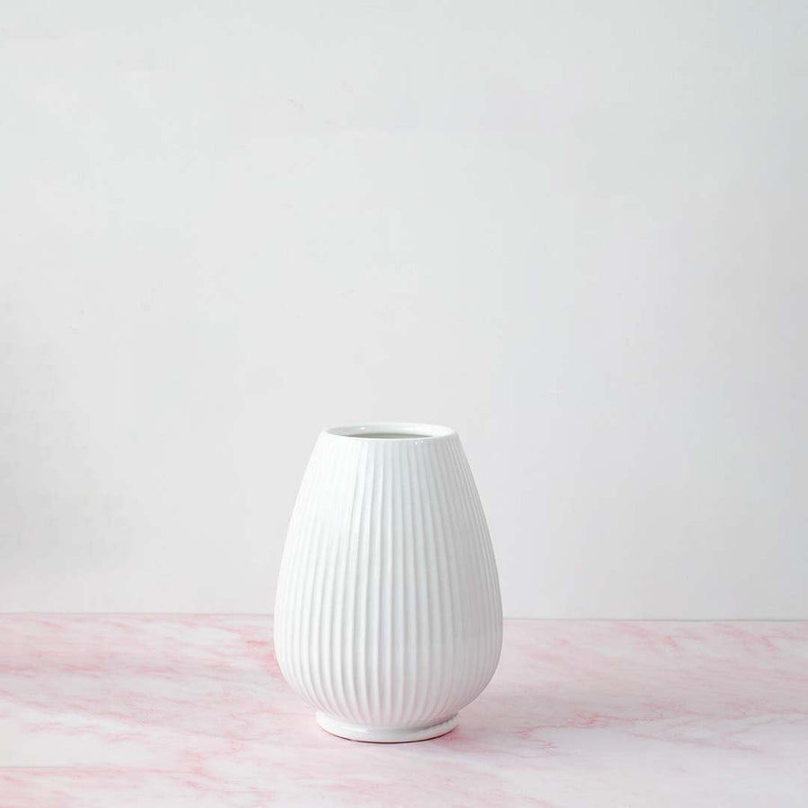White ribbed ceramic vase with natural lighting on white background creating a soft shadow and minimalist artistic vibe.