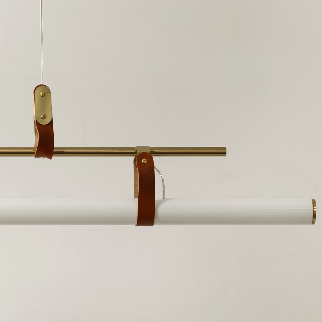 Detailed closeup of gold linear pendant light showing metallic frame leather straps and smooth white diffuser tube.