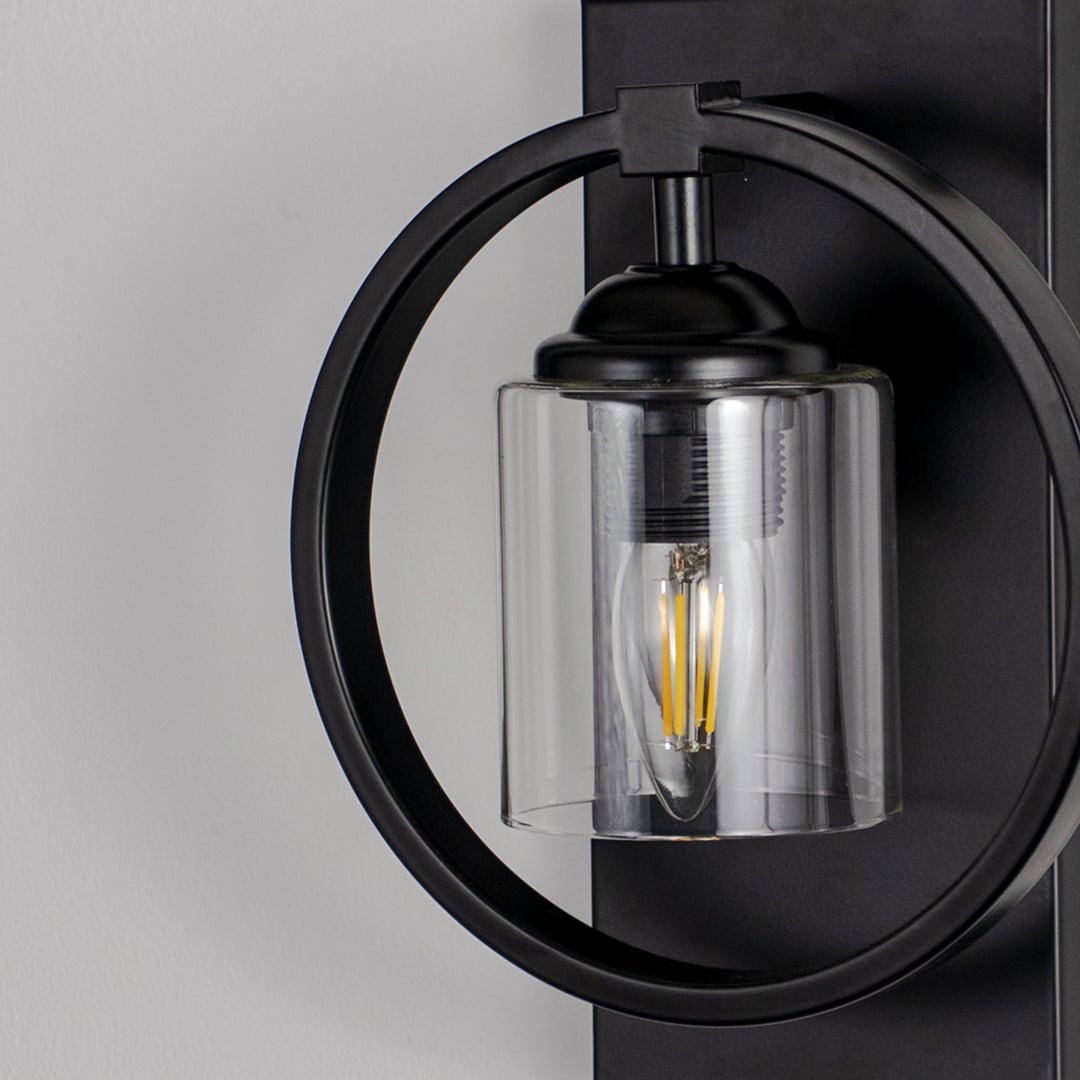 Closeup of round black wall light with cylindrical glass shade highlighting exposed filament bulb and sleek metal frame.