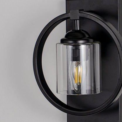 Closeup of round black wall light with cylindrical glass shade highlighting exposed filament bulb and sleek metal frame.