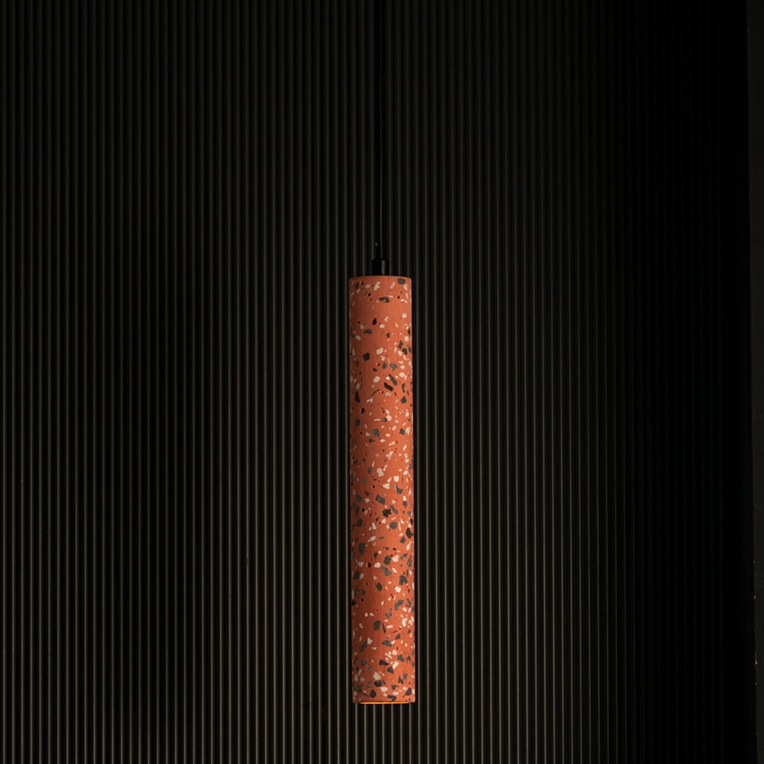 Terracotta cylinder pendant light turned on glowing warmly with terrazzo finish and brass detailing against dark background.