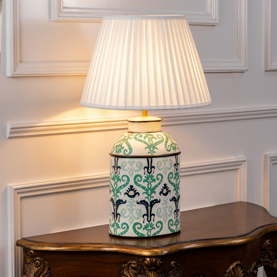Table lamp with a white pleated shade and mint green ceramic base decorated with traditional floral patterns on a wooden surface.