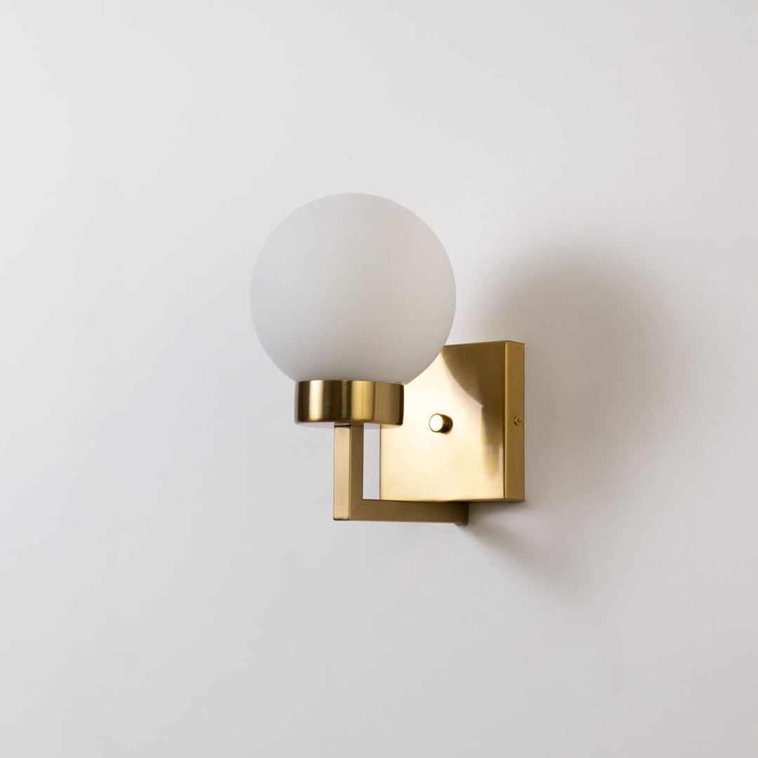 Gold metal wall light with frosted glass globe shade on bright white background with light off highlighting its sleek modern form.