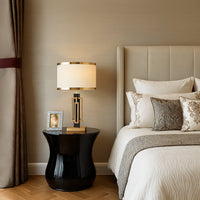 Table lamp with black and gold geometric base and drum shade placed on a glossy black side table creating warm ambient lighting in an elegant bedroom decor.