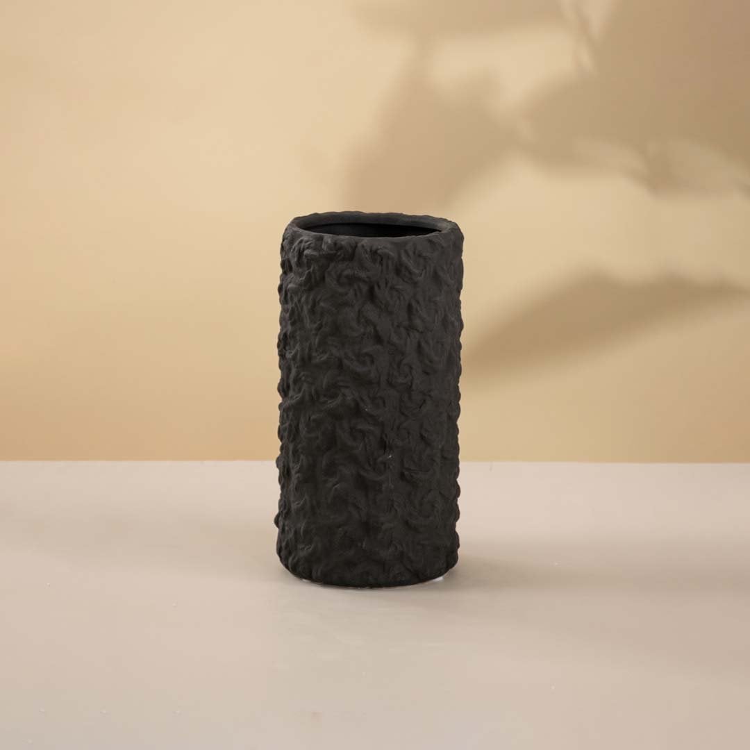 Matte black cylindrical vase with rough handcrafted texture holding white florals against white background placed on a classic wooden surface.