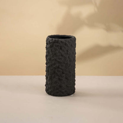Matte black cylindrical vase with rough handcrafted texture holding white florals against white background placed on a classic wooden surface.