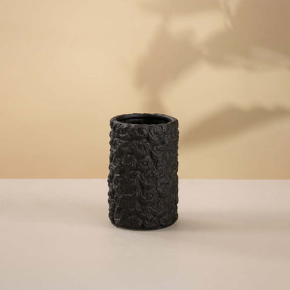 Black handcrafted ceramic vase with cylindrical shape and textured detailing against neutral white backdrop.