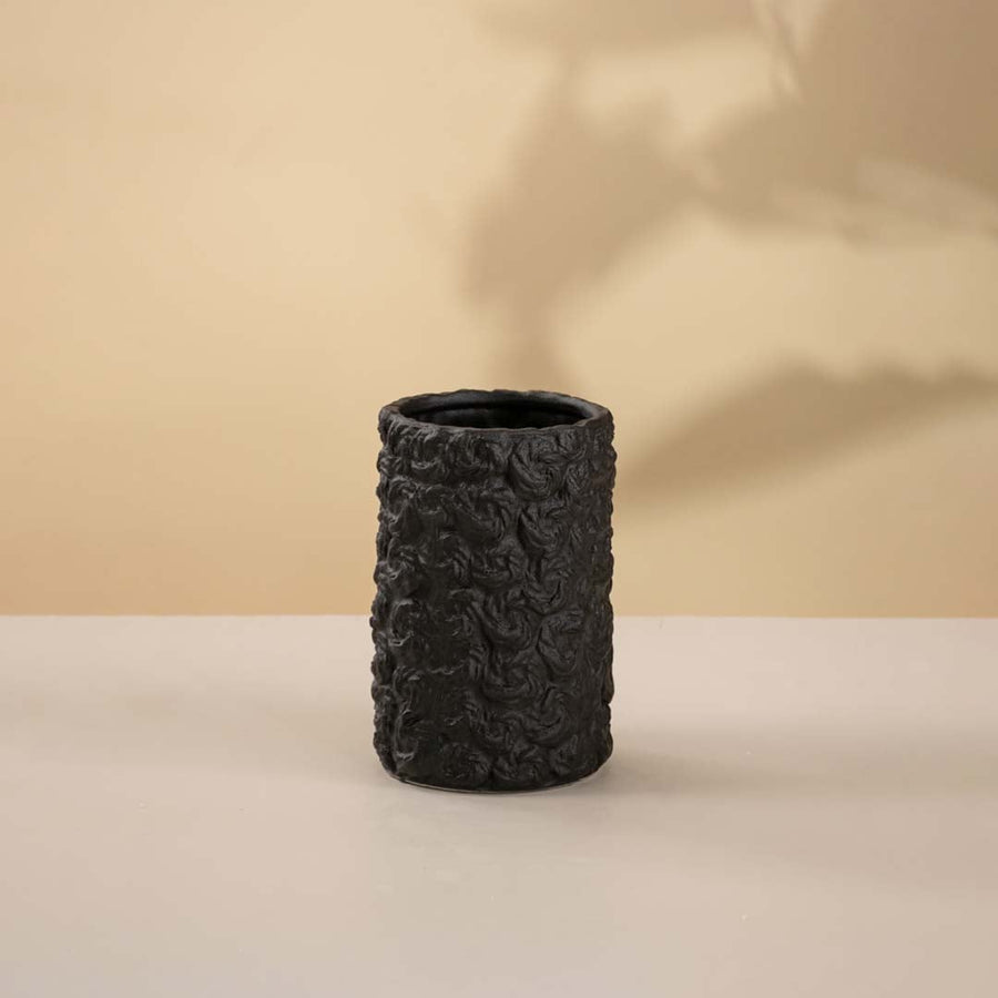 Black handcrafted ceramic vase with cylindrical shape and textured detailing against neutral white backdrop.
