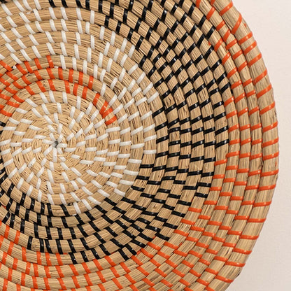 Closeup view of circular woven wall decor with layered spiral pattern in beige black and orange tones for textured wall styling.