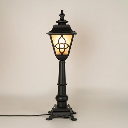 A traditional outdoor pillar lamp displayed on a white background with the light on showing its frosted glass panels ornate metal pattern and tall sculpted post design.