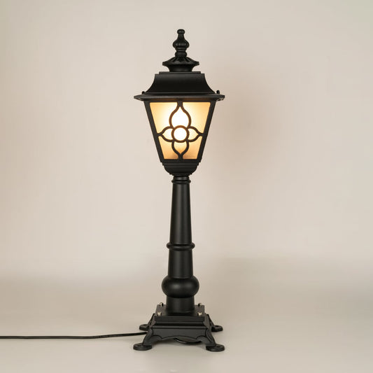 A traditional outdoor pillar lamp displayed on a white background with the light on showing its frosted glass panels ornate metal pattern and tall sculpted post design.