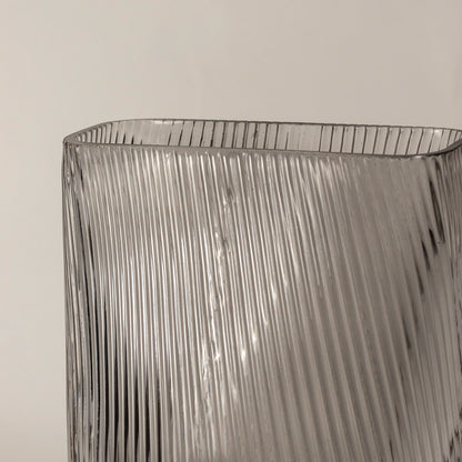 Tall clear ribbed glass vase closeup with vertical texture placed on wooden surface against neutral wall background enhancing modern decorative styling.