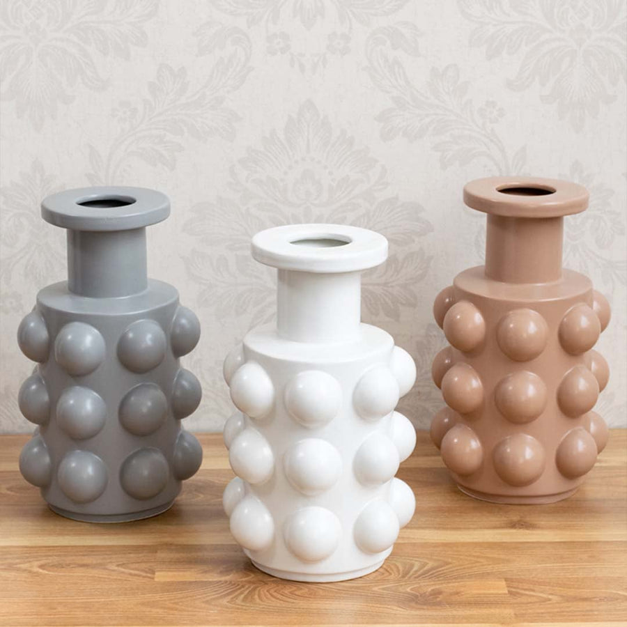 Pair of modern white ceramic vases with circular raised detailing placed on a wooden shelf with books and greenery.