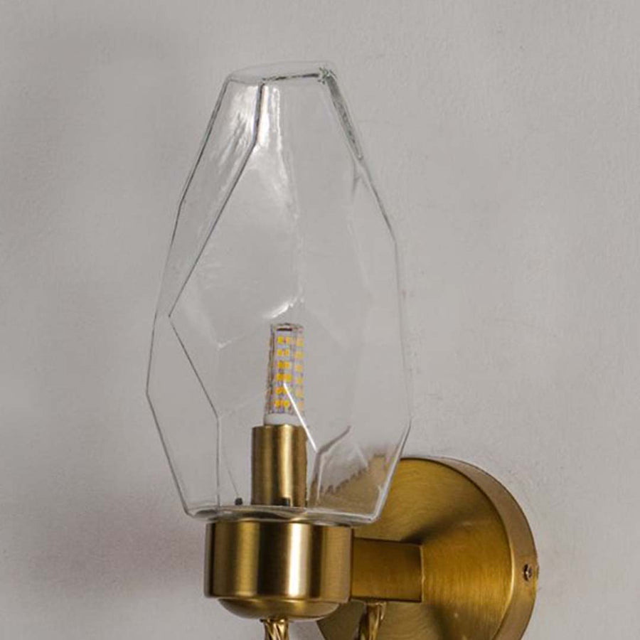 Closeup of wall lamp with geometric clear glass shade and gold finish base showcasing elegant luxury wall mounted lighting design.