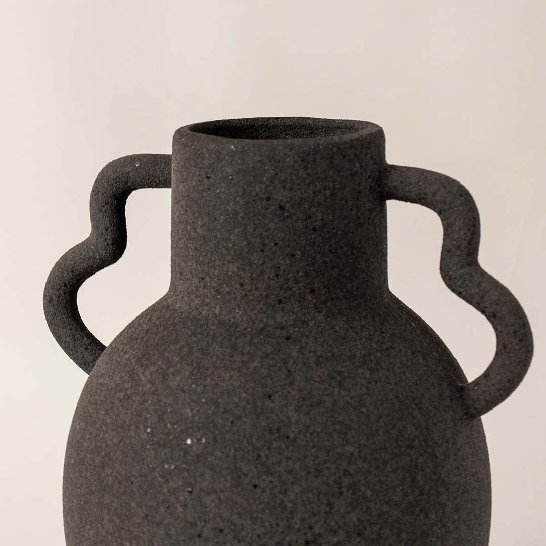 Dark textured ceramic vase with dual side handles placed on white surface against neutral background showcasing rustic modern home decor styling.