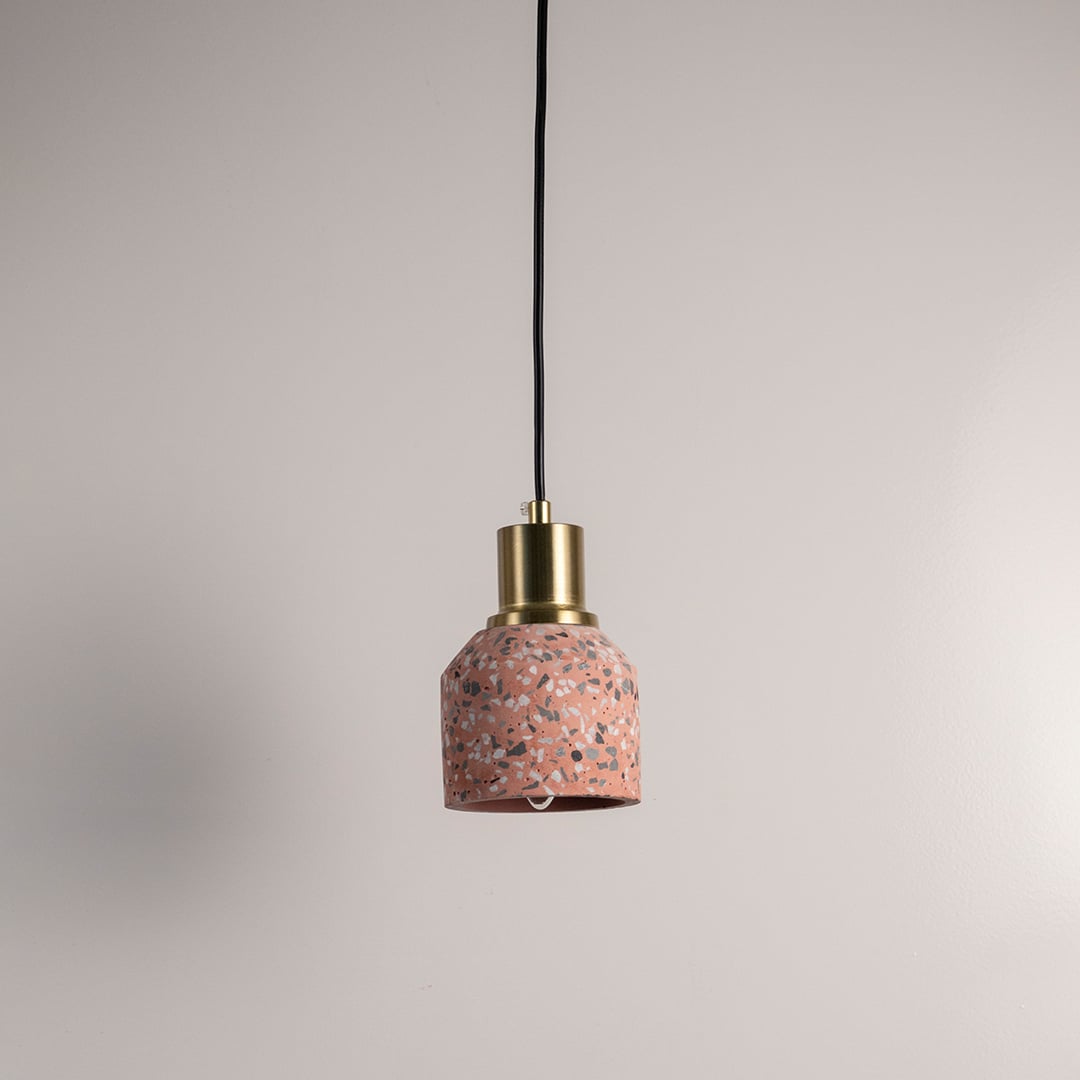 Pink terrazzo pendant light with brass top and black cord displayed against white wall.