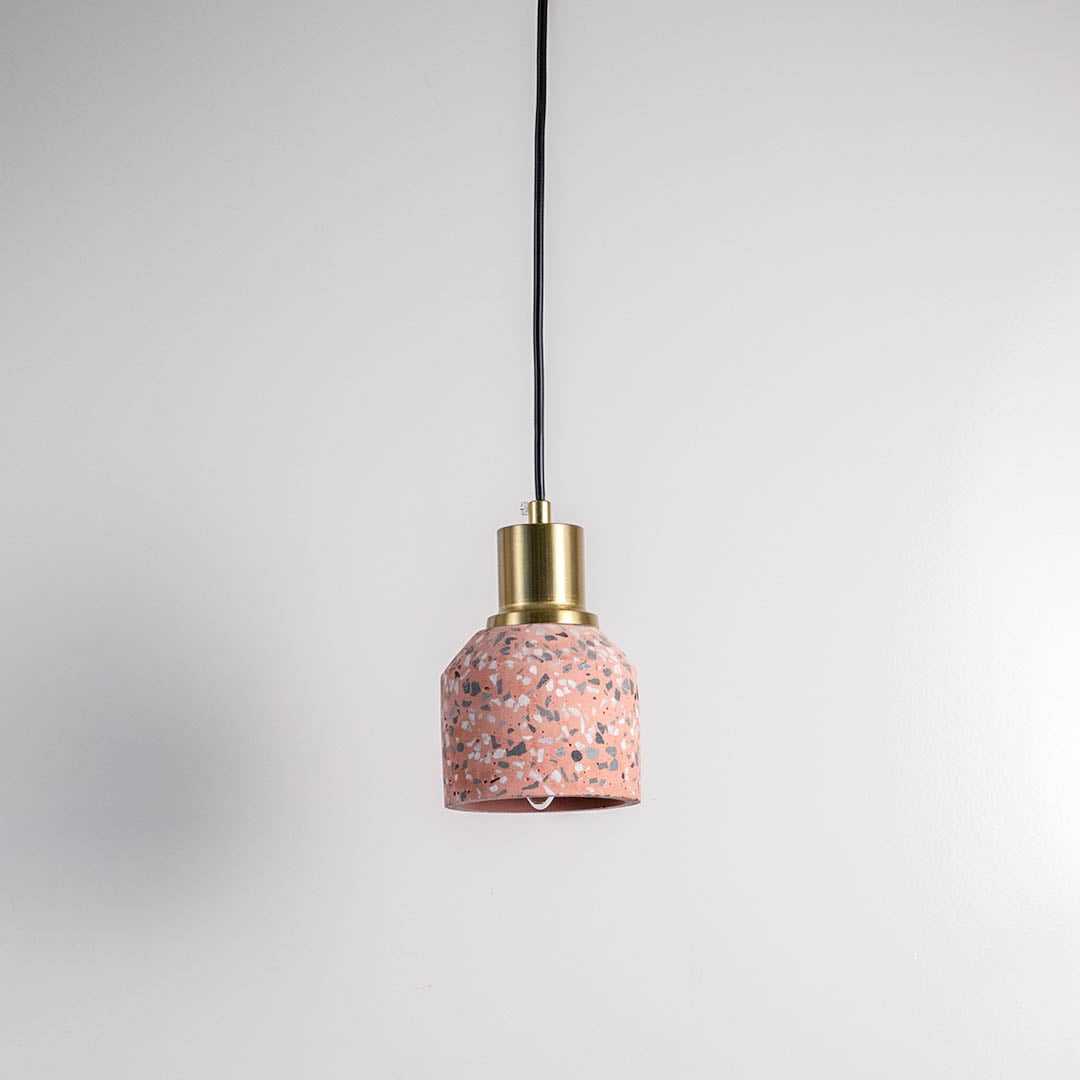 Pink terrazzo pendant light with brass top and black cord displayed against white wall in modern minimal look.