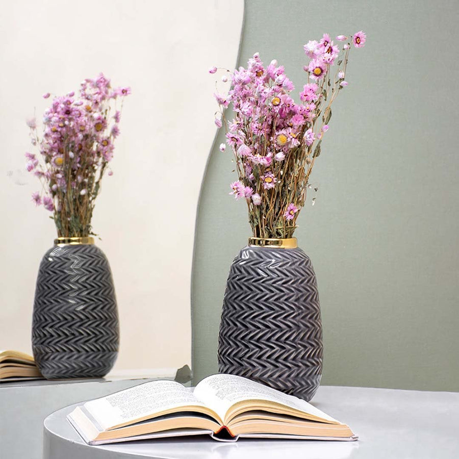 A decorative grey vase with a zigzag pattern filled with pink dried flowers placed beside an open book on a reflective table creating a soft elegant lifestyle scene.
