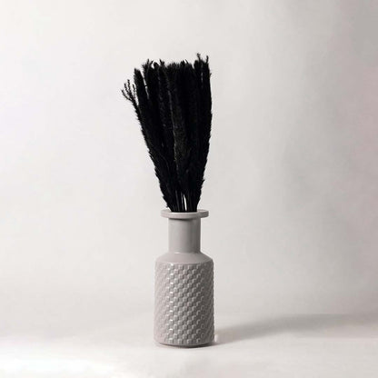A decorative vase with a textured pattern holding tall black dried stems placed against a clean white background showing a minimal and modern home decor accent.