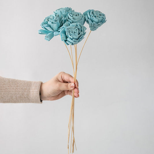 Hand holding a bundle of turquoise blue handmade sola wood flower stems for modern coastal home vase decor on white backdrop.