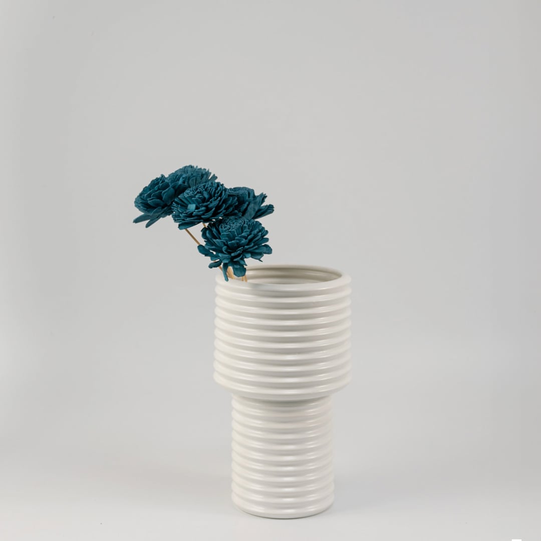 Blue artificial flowers in glass vase placed near curtain and green plant on white table for elegant home styling.