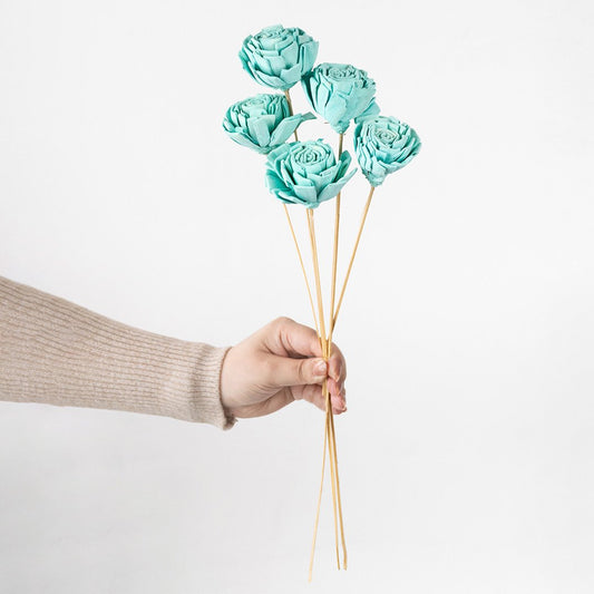 Hand holding a bundle of turquoise blue handmade sola wood flower stems for modern coastal home vase decor on white backdrop.