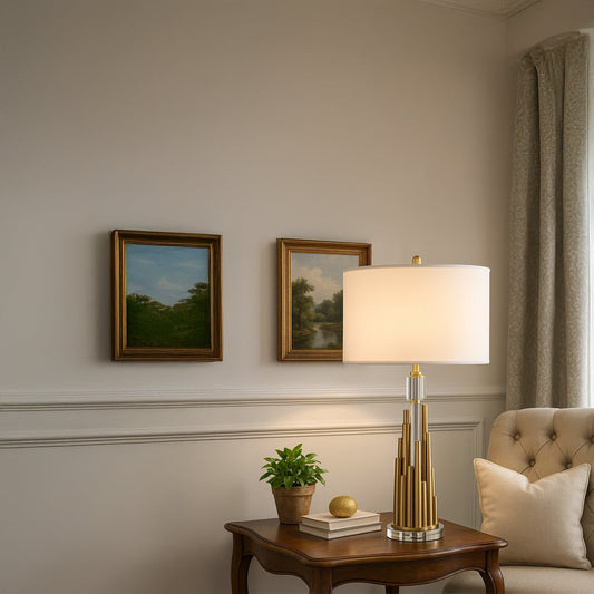 Gold metal table lamp with tiered cylindrical base and white drum shade placed on a wooden side table creating warm ambient lighting in an elegant living space.