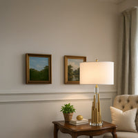 Gold metal table lamp with tiered cylindrical base and white drum shade placed on a wooden side table creating warm ambient lighting in an elegant living space.