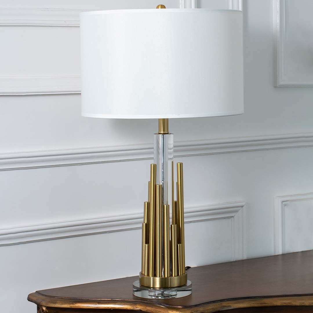 Gold metal table lamp with white drum shade and sculpted tiered base displayed on a white background perfect for modern home styling and console table decor.