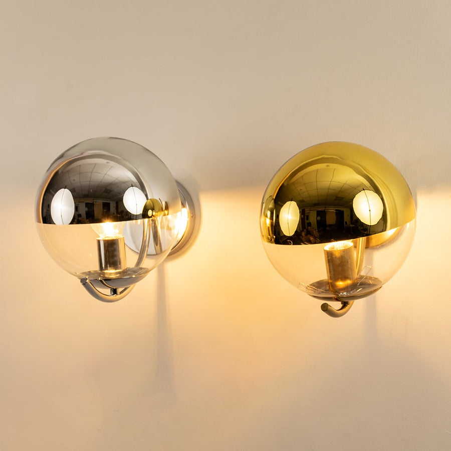 A duo of metal wall lights in chrome and gold finishes glowing warmly against a clean white wall creating reflective highlights and modern luxurious décor.
