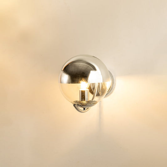 A chrome metal wall light with a spherical shade glowing on a beige wall creating reflective highlights and adding sleek modern décor.