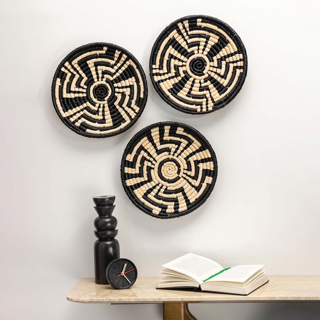 Set of three black and beige seagrass wall plates with geometric maze patterns arranged on a white wall above a marble table.