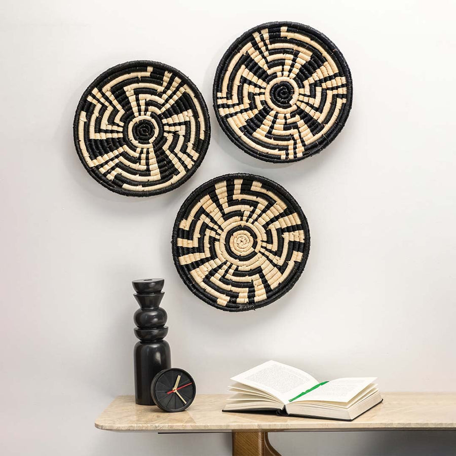 Set of three black and beige seagrass wall plates with geometric maze patterns arranged on a white wall above a marble table.