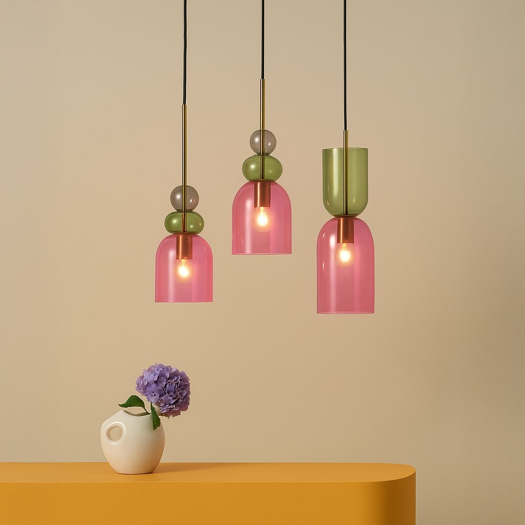 Modern pendant lights with pink and green glass shades hanging above a yellow table with a white vase and flower.