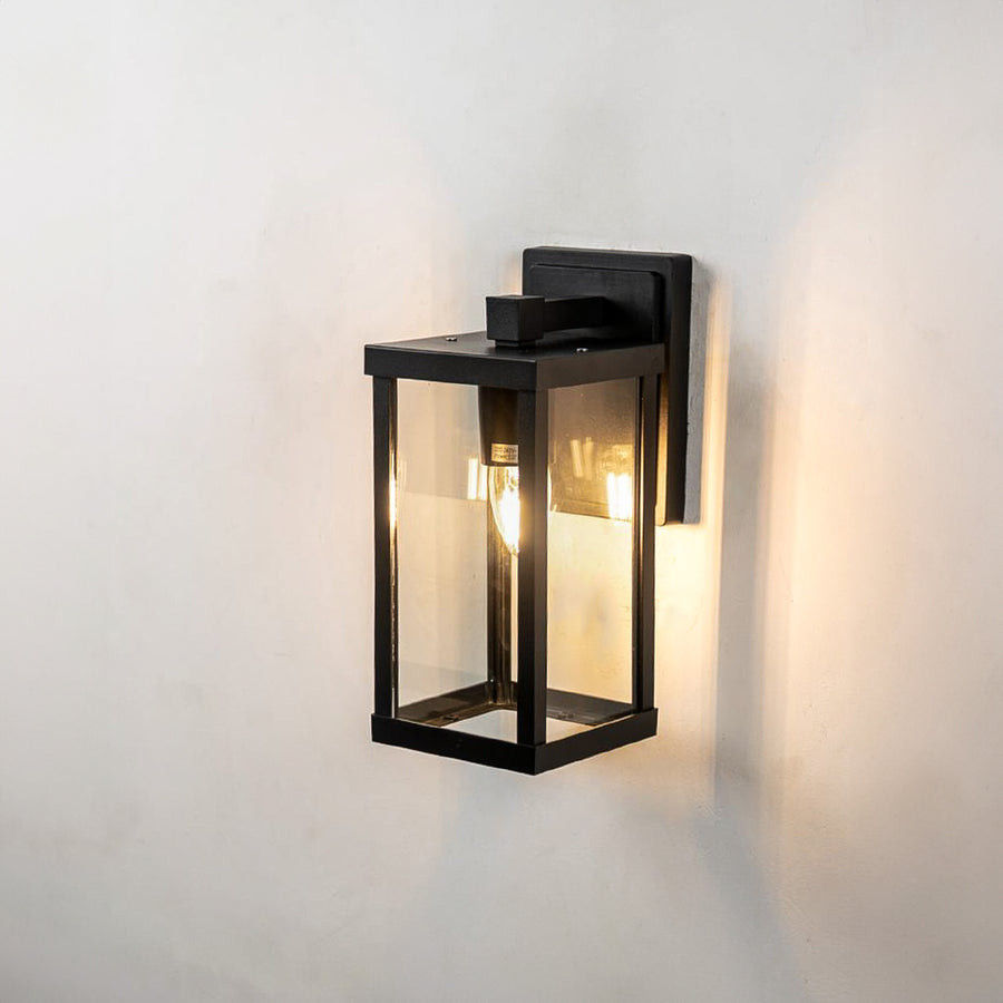 Dual bulb black metal outdoor sconce with clear glass casing casting a warm light reflection on white background wall.